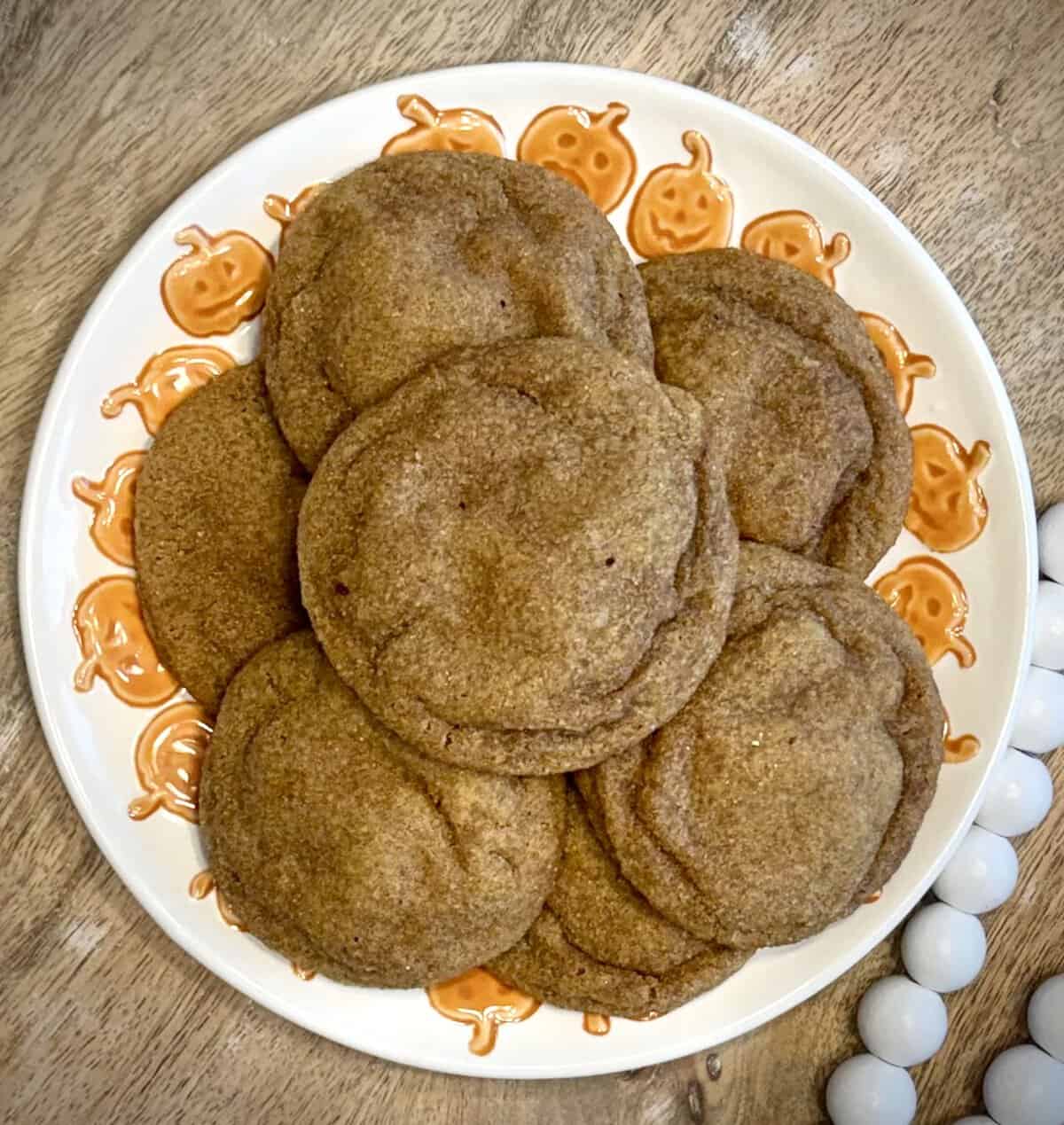 A plate of cookies sits on a wooden table, decorated with orange pumpkin faces around the plate’s rim.