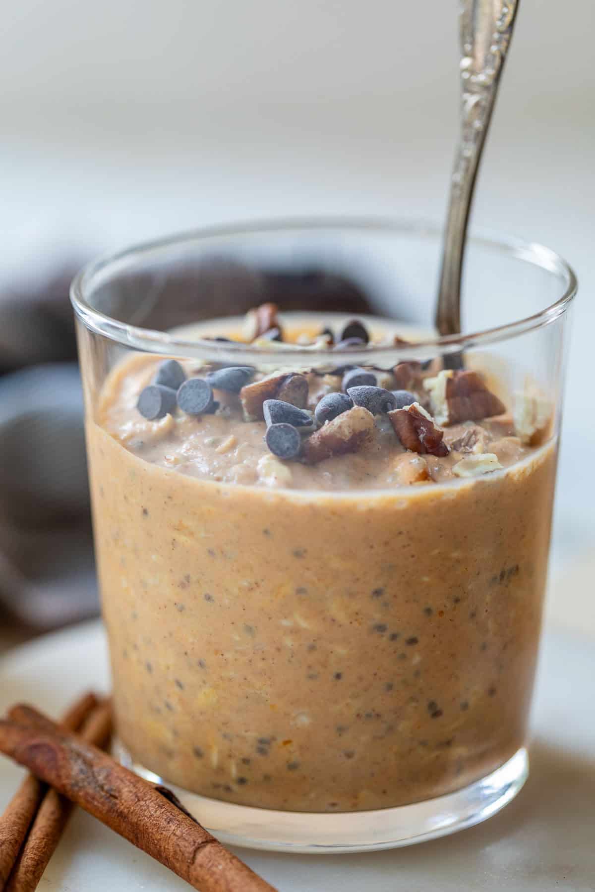 A glass of creamy pumpkin overnight oats topped with chocolate chips and nuts, with a spoon and cinnamon sticks beside it.