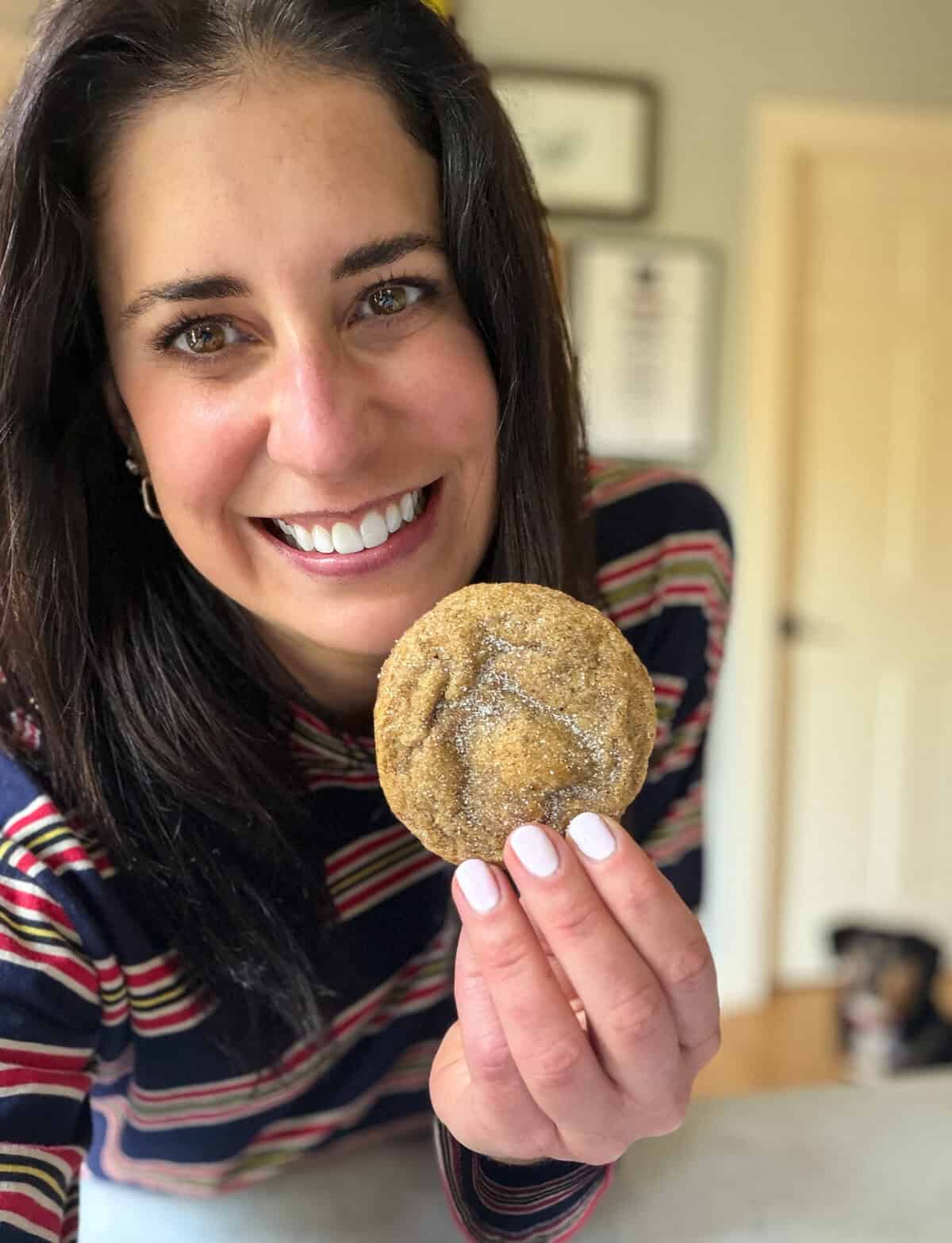 Woman smiling and holding a sugar-coated cookie close to the camera in a bright kitchen.