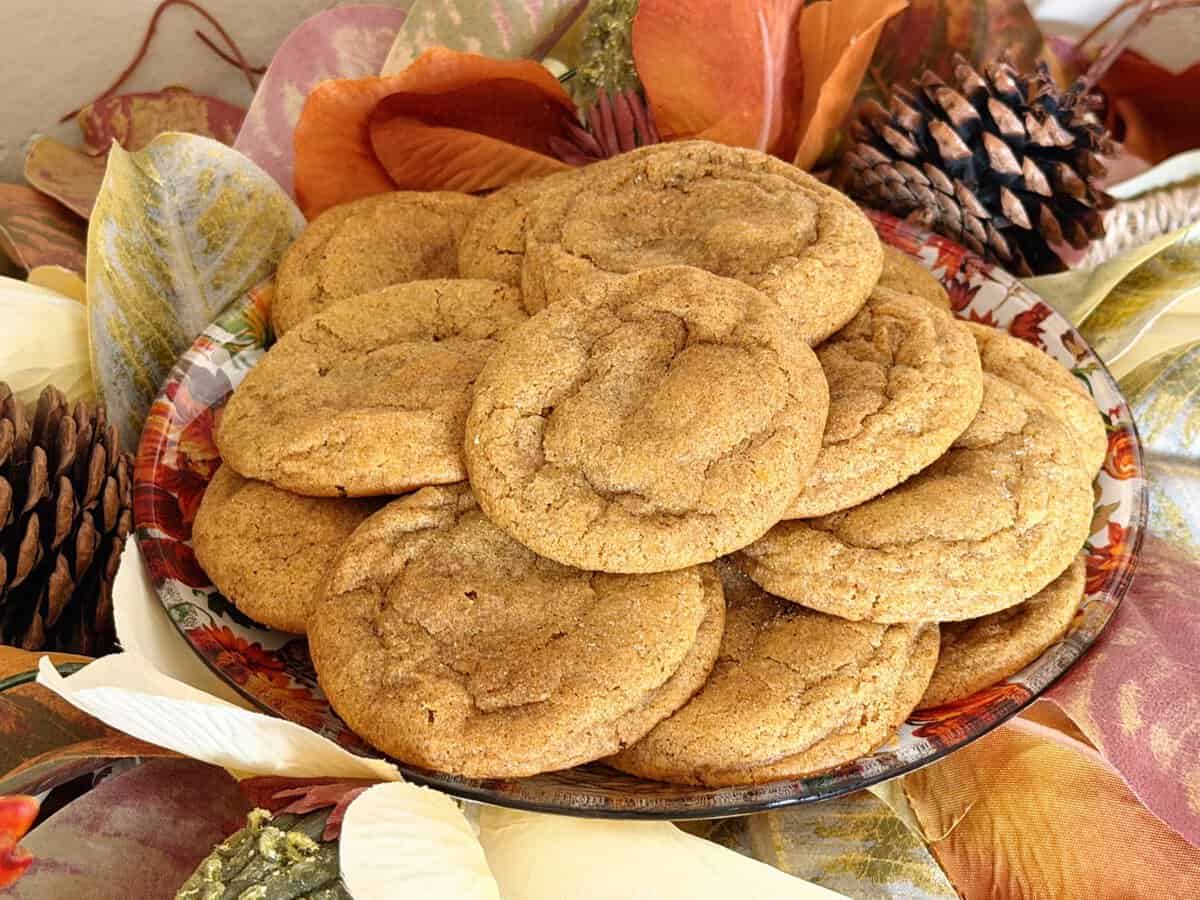 A plate of brown cookies sits on autumn leaves and pinecones, creating a festive, cozy fall atmosphere.