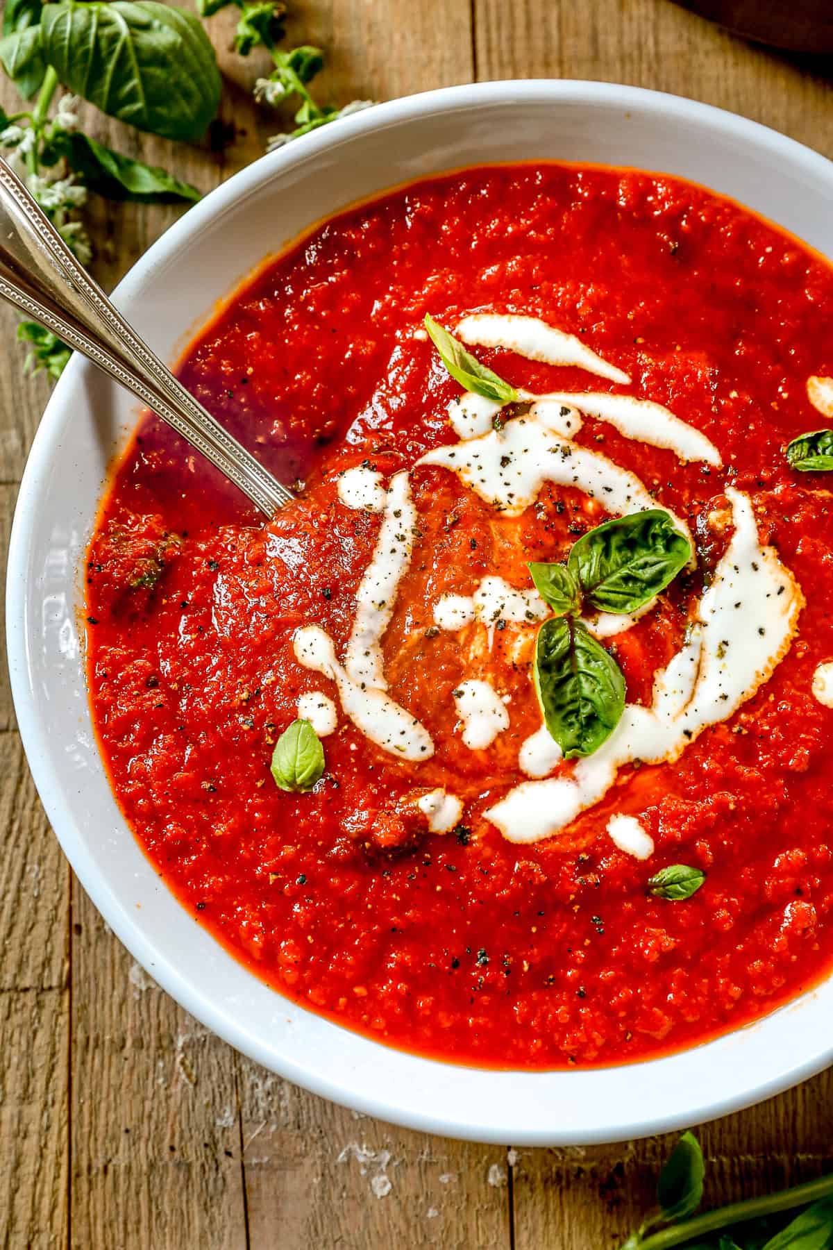 Bowl of roasted red pepper soup with cream swirls, basil leaves, and a spoon, on a wooden table.