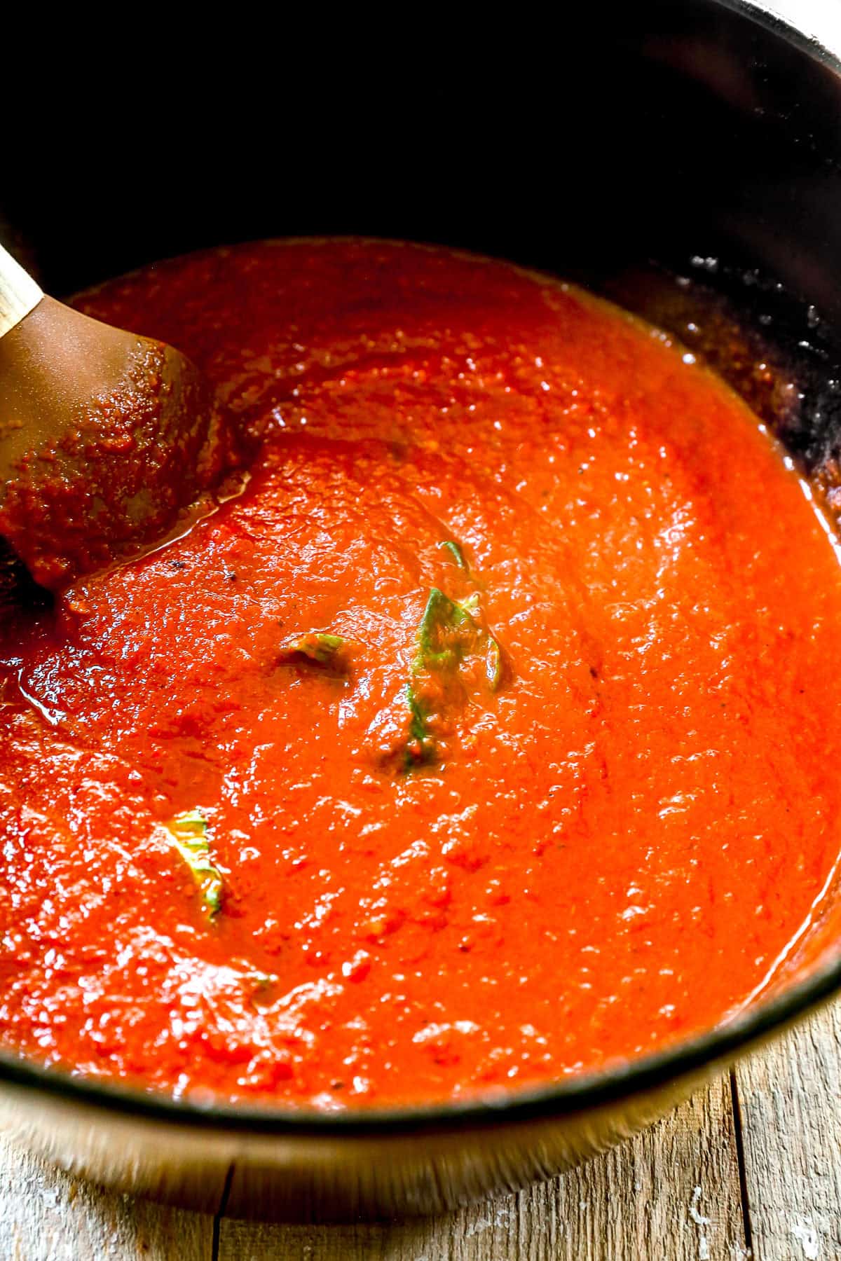 Roasted red pepper soup with basil leaves being stirred in a black pot using a wooden spoon on a wooden surface.