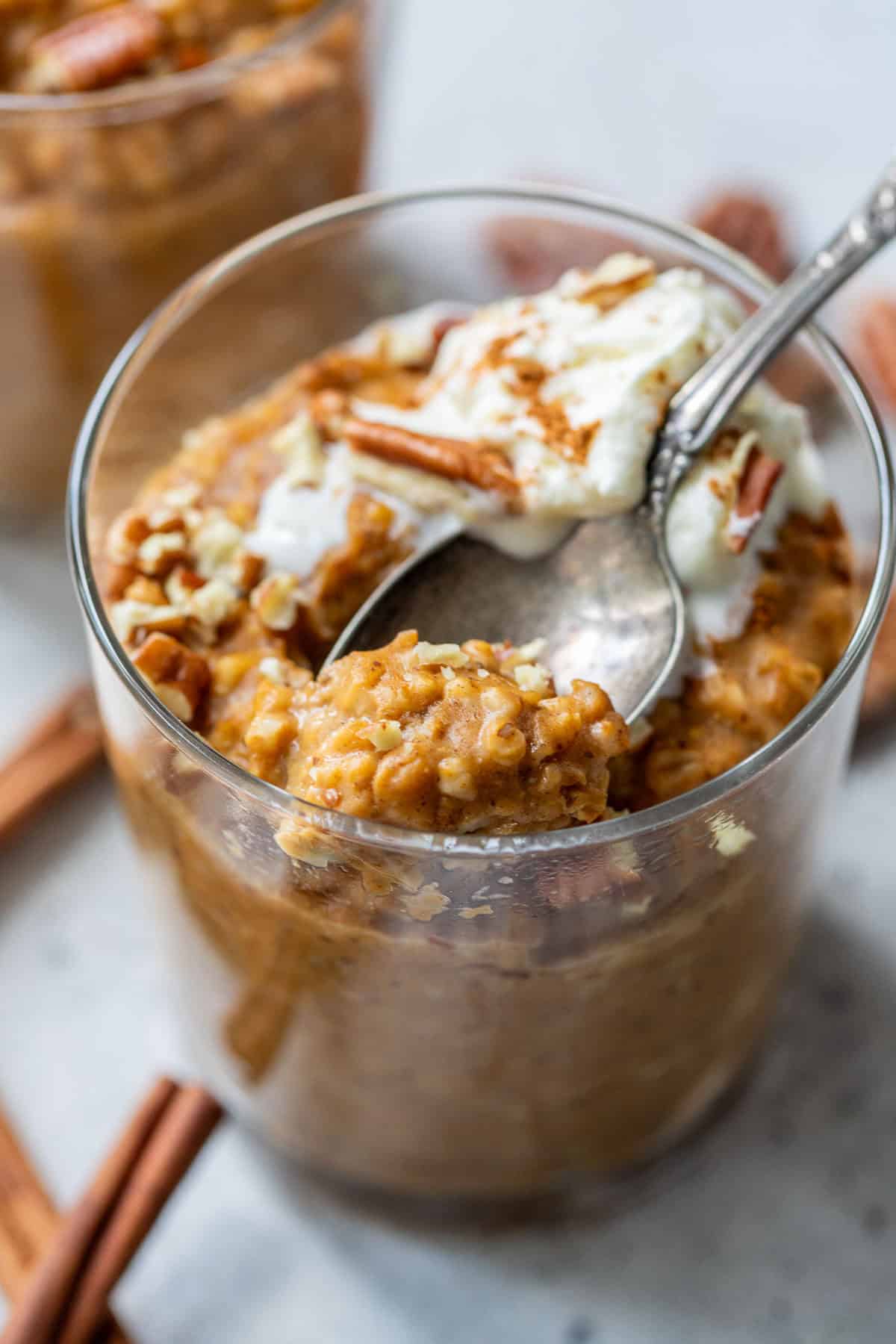 Glass of creamy slow cooker pumpkin oatmeal topped with whipped cream, pecans, and cinnamon sticks, with a spoon scooping some out.