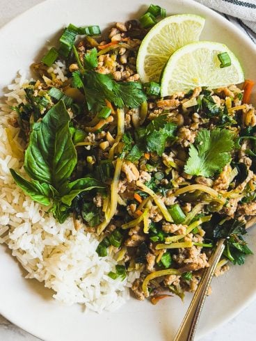 A plate of white rice and ground chicken stir fry with herbs, garnished with lime slices, basil, and cilantro.