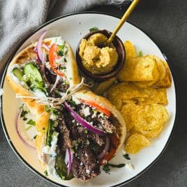 A plate with two gyros, potato chips, a bowl of hummus, and a beige napkin beside the plate.