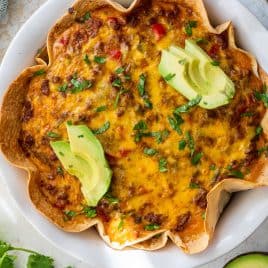 Baked taco pie with melted cheese, chopped herbs, and sliced avocado on top, served on a white plate.