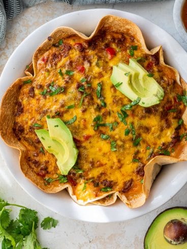 Baked taco pie with melted cheese, chopped herbs, and sliced avocado on top, served on a white plate.