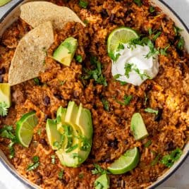 One-pot taco rice with beans, avocado, lime, tortilla chips, sour cream, and cilantro in a round pan.