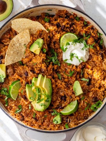One-pot taco rice with beans, avocado, lime, tortilla chips, sour cream, and cilantro in a round pan.