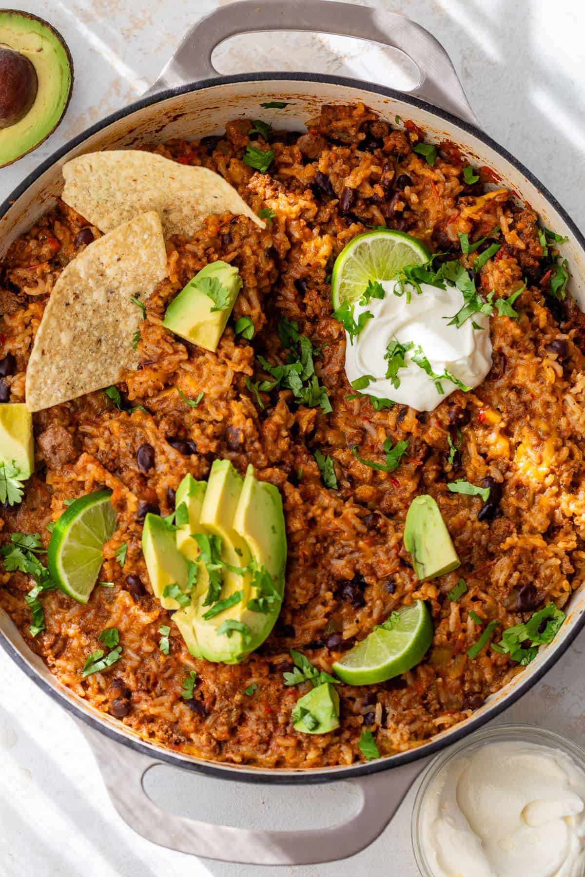 One-pot taco rice with beans, avocado, lime, tortilla chips, sour cream, and cilantro in a round pan.