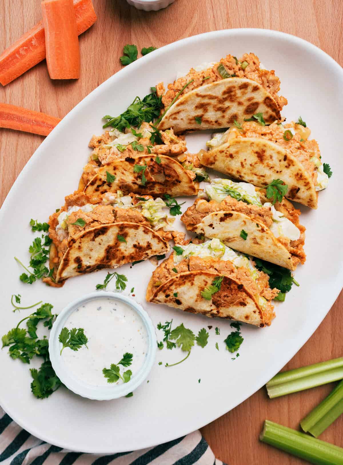 A plate of buffalo chicken smash tacos with creamy filling, garnished with cilantro, served with dip, celery, and carrot sticks.