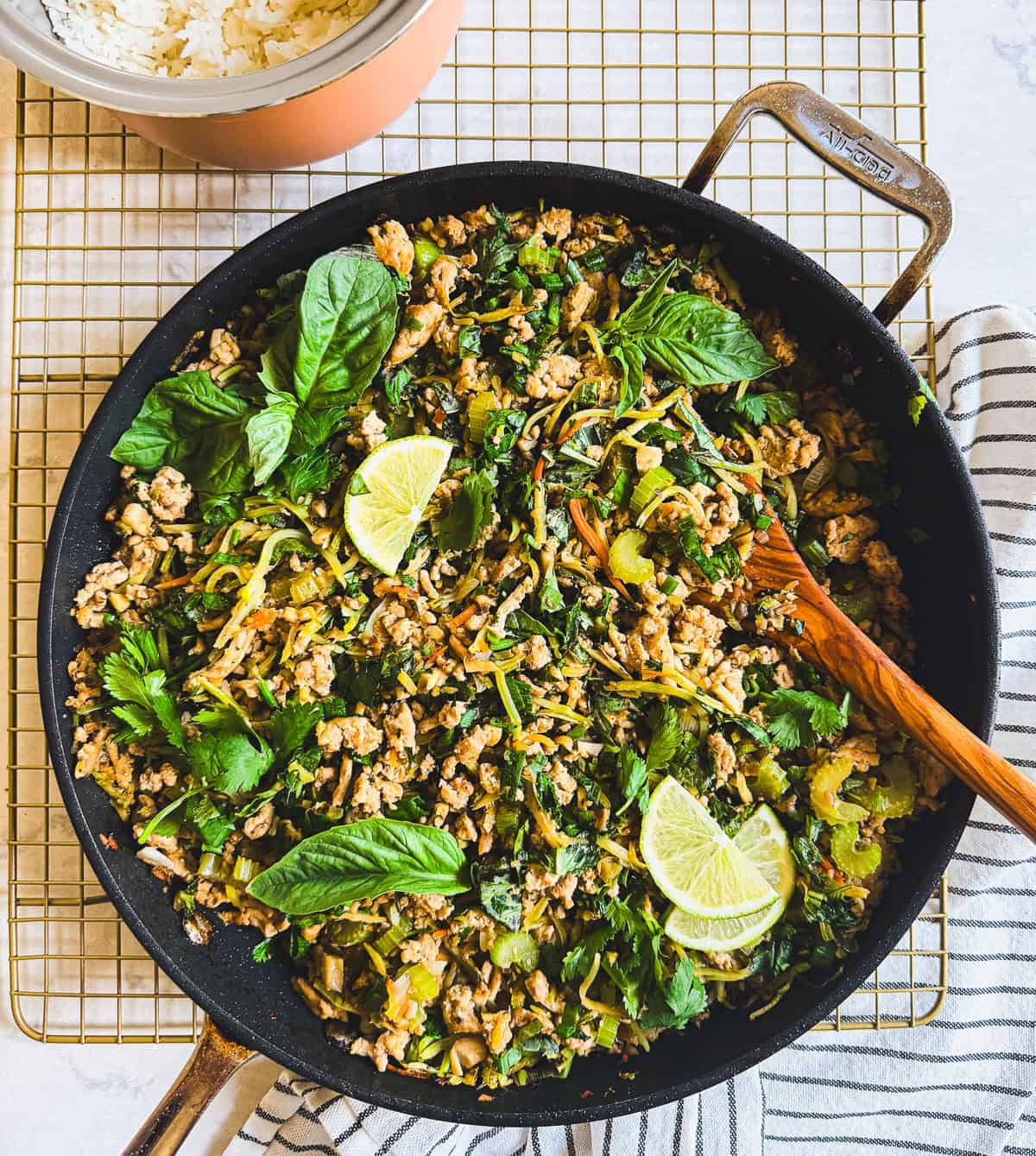 A skillet of ground chicken stir fry with herbs, greens, and lime wedges, next to a bowl of rice on a cooling rack.