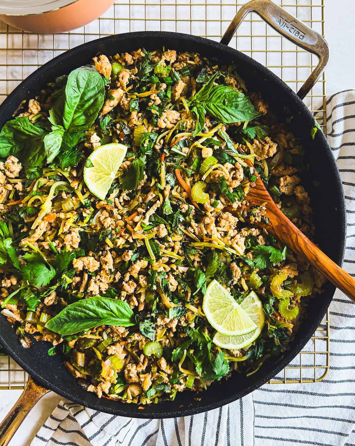A skillet of ground chicken stir-fry with greens, herbs, and lime wedges, with a wooden spoon and striped towel nearby.
