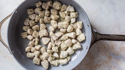 Cubed, seasoned chicken cooking in a large gray skillet on a light, textured surface.