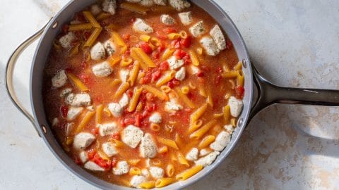 A skillet with penne pasta, diced chicken, and tomatoes in a red sauce, ready to be cooked.