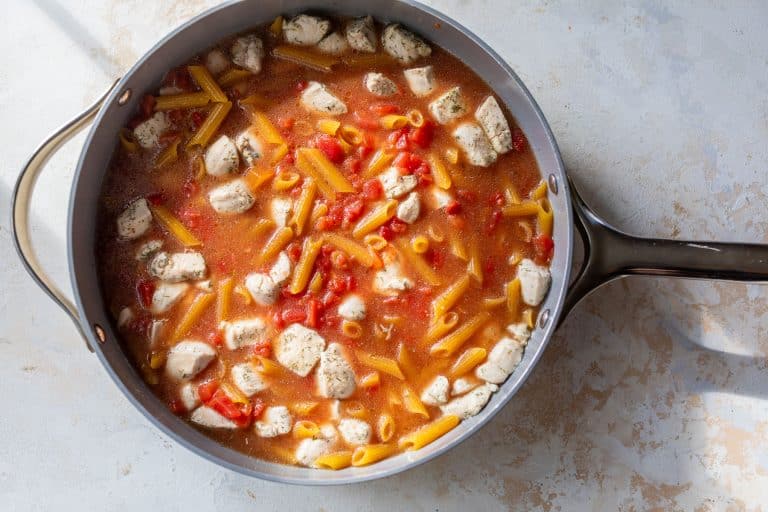 A skillet with penne pasta, diced chicken, and tomatoes in a red sauce, ready to be cooked.