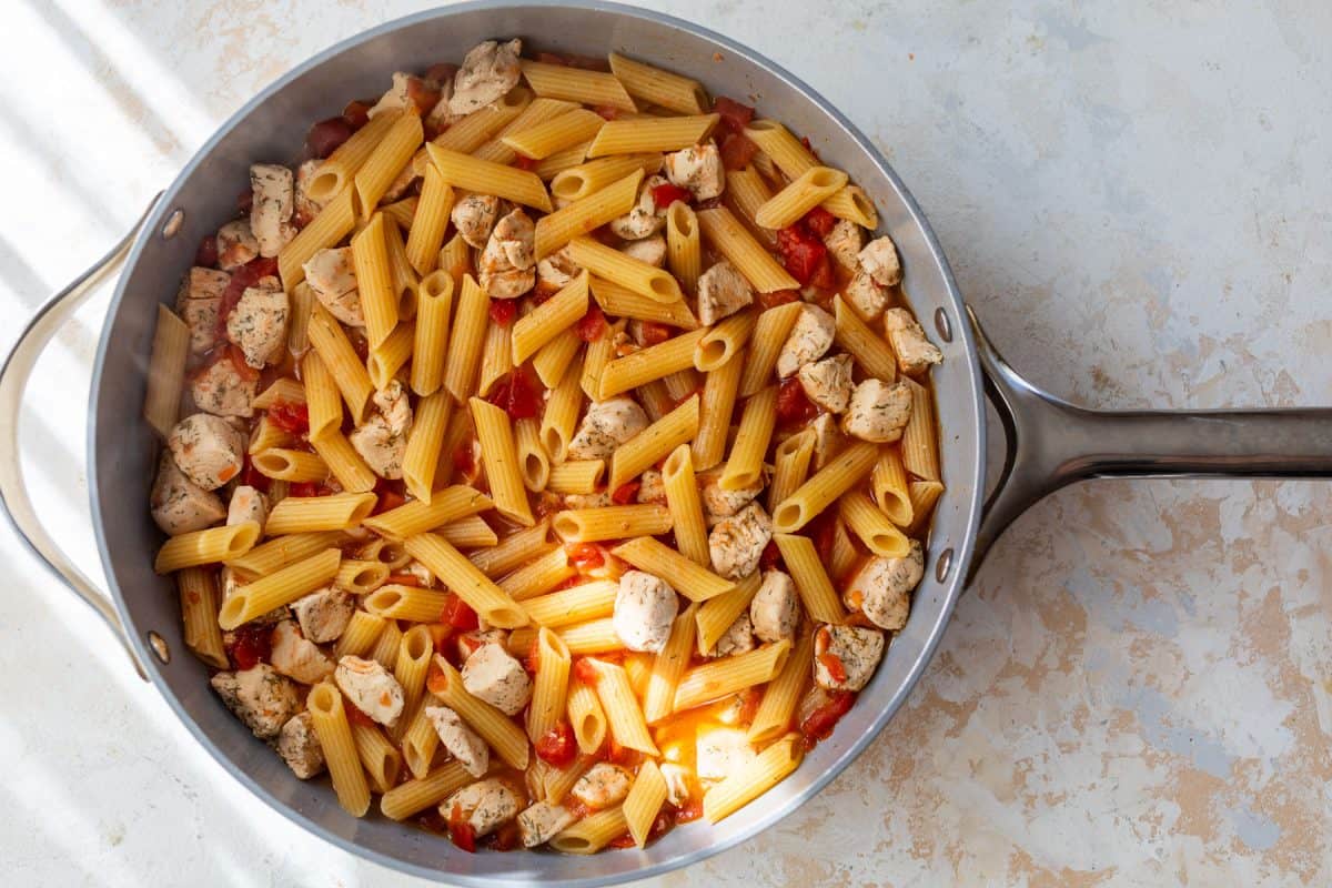Pan of cooked penne pasta with diced chicken and tomatoes on a light textured surface.