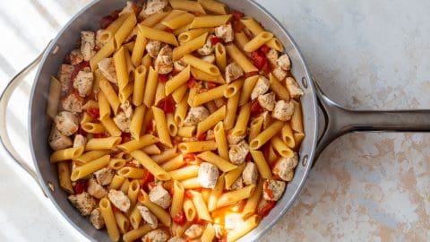 Pan of cooked penne pasta with diced chicken and tomatoes on a light textured surface.