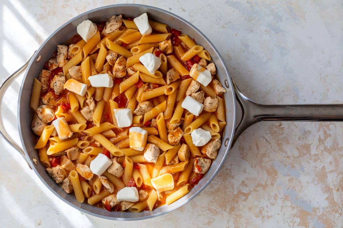 A skillet with penne pasta, chicken, buffalo sauce, and dollops of cream cheese on a light countertop.