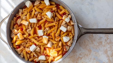 A skillet with penne pasta, chicken, buffalo sauce, and dollops of cream cheese on a light countertop.