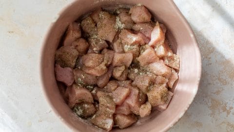 Raw, seasoned chicken pieces in a pink bowl on a light-colored surface, ready to be cooked.