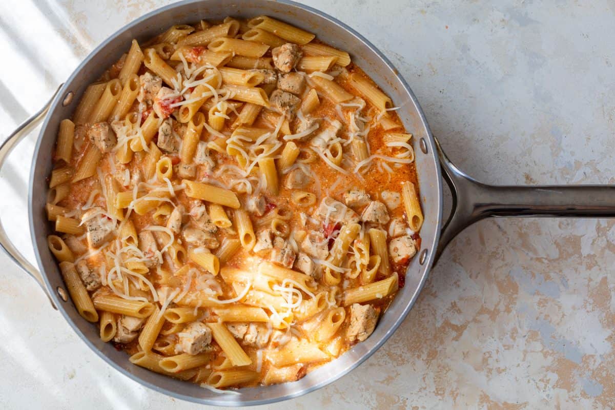 A skillet of creamy buffalo pasta with chicken chunks and shredded cheese on top.