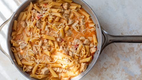 A skillet of creamy buffalo pasta with chicken chunks and shredded cheese on top.
