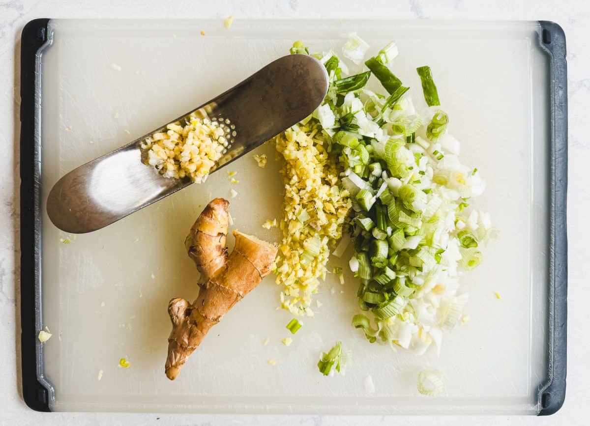 Chopped ginger, green onions, and a ginger root on a cutting board with a metal grater.