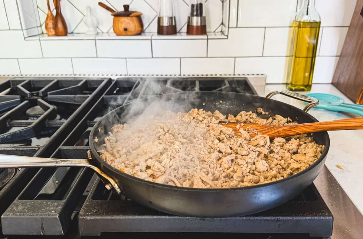 A pan of ground chicken cooking on a stovetop with a wooden spoon; steam is rising from the pan.