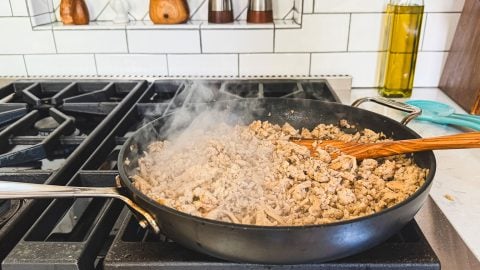 A pan of ground chicken cooking on a stovetop with a wooden spoon; steam is rising from the pan.