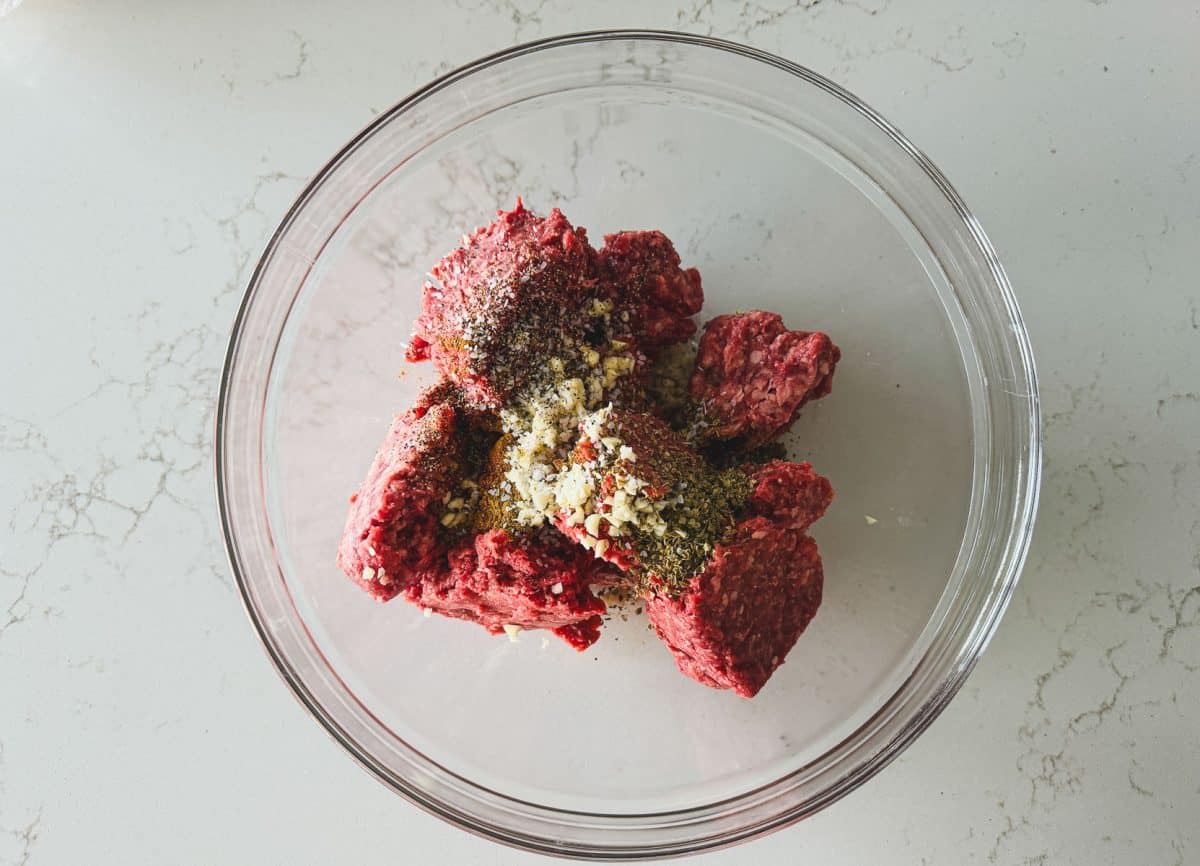 Ground beef and lamb in a clear bowl topped with various spices and minced garlic, on a light marble countertop.