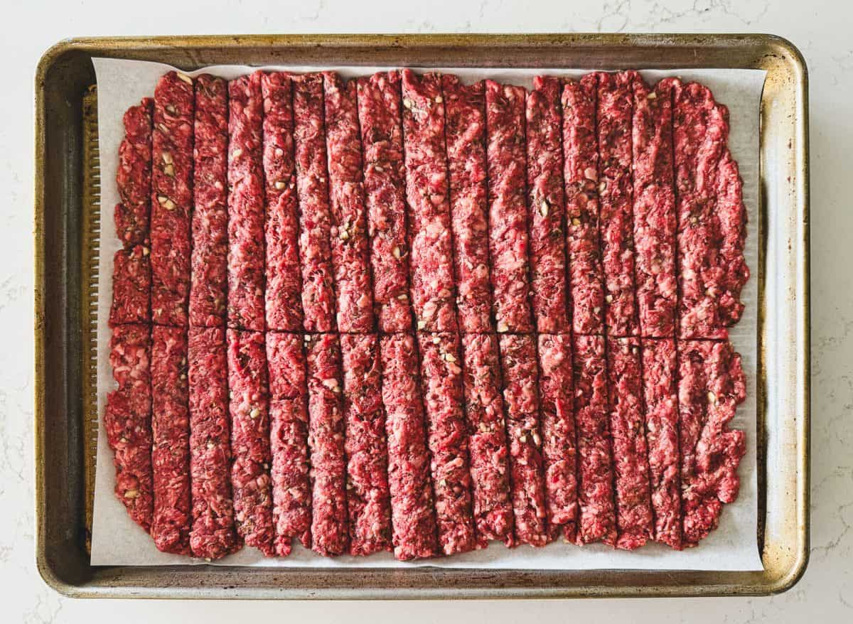 Raw ground beef pressed flat and cut into uniform strips on a parchment-lined baking sheet for gyro recipe.