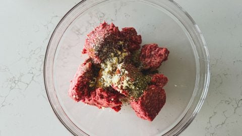 Ground beef and lamb in a clear bowl topped with various spices and minced garlic, on a light marble countertop.