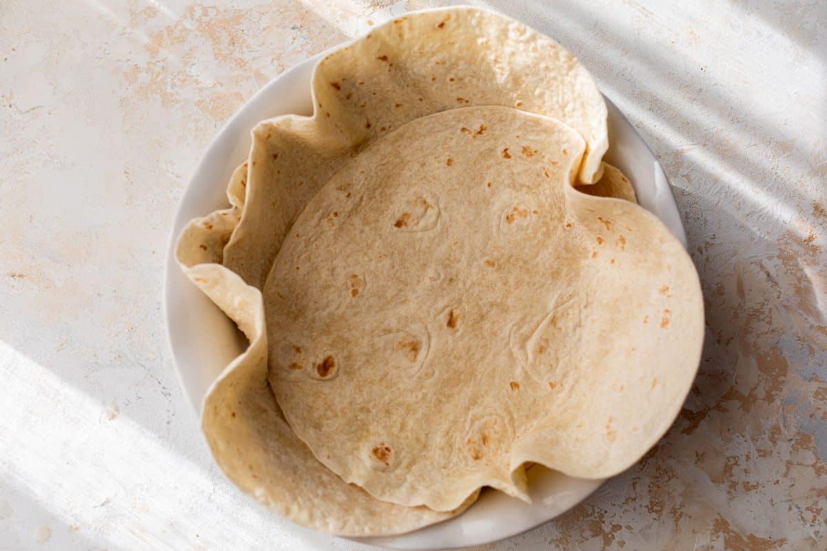 A white plate with several flour tortillas, slightly overlapping, on a light textured surface.