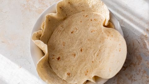A white plate with several flour tortillas, slightly overlapping, on a light textured surface.
