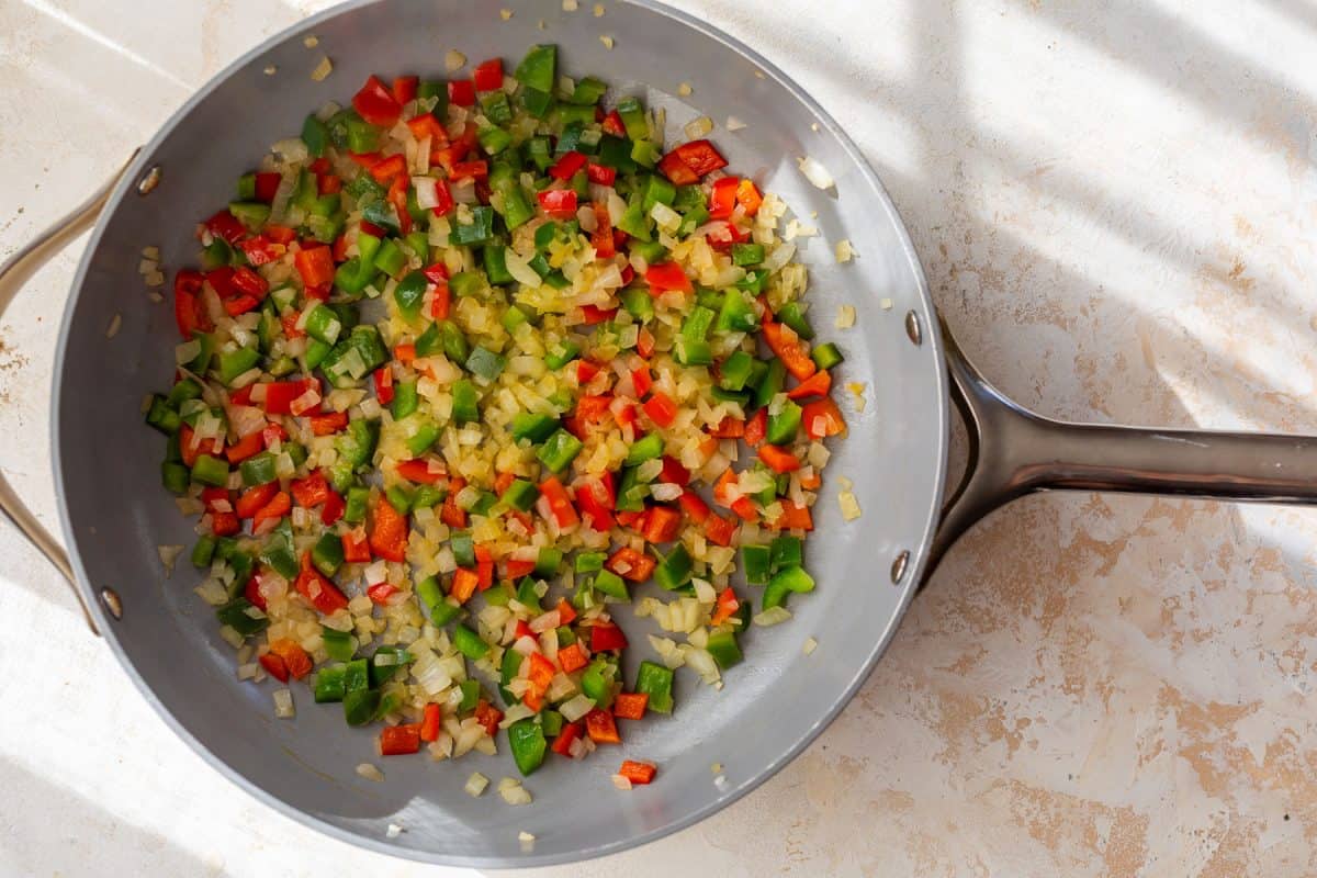 Chopped onions, red, and green bell peppers cooking in a skillet on a light-colored surface.