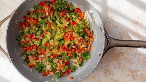Chopped onions, red, and green bell peppers cooking in a skillet on a light-colored surface.