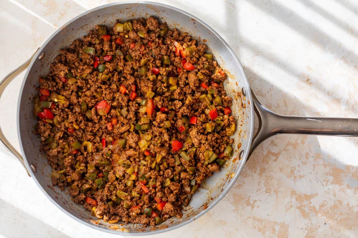 A skillet filled with seasoned ground beef, diced red and green bell peppers on a light countertop.