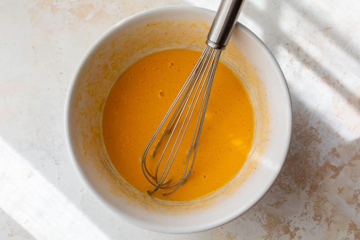 A metal whisk in a white bowl with beaten eggs on a light-colored surface, lit by natural sunlight.