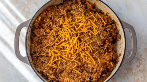 A large skillet of cheesy taco rice, beans, and ground beef, topped with shredded cheddar cheese on a light surface.