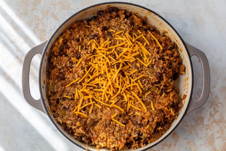 A large skillet of cheesy taco rice, beans, and ground beef, topped with shredded cheddar cheese on a light surface.