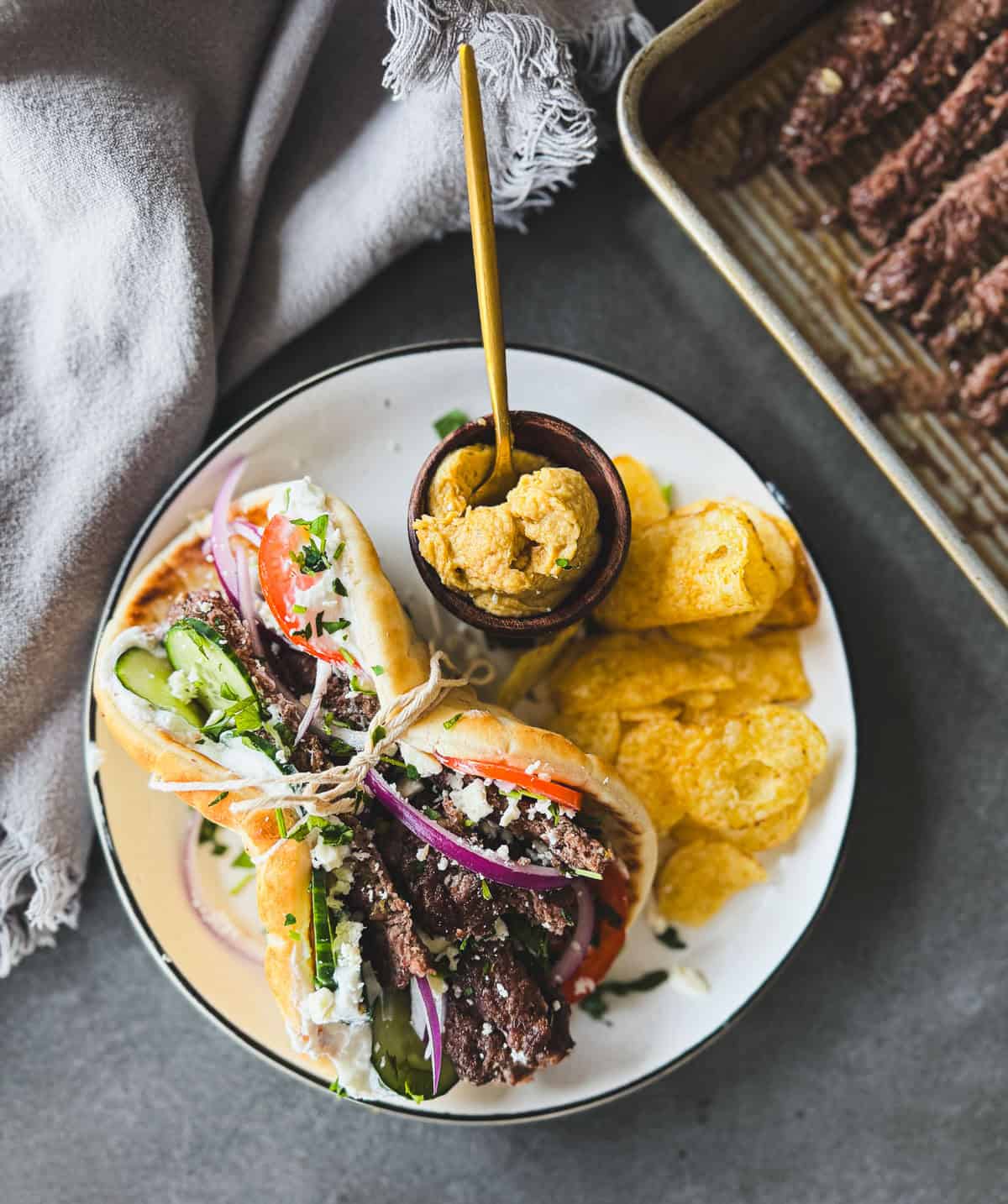 Gyro sandwich with veggies and feta, served with chips, mustard dip, on a white plate next to a baking tray.