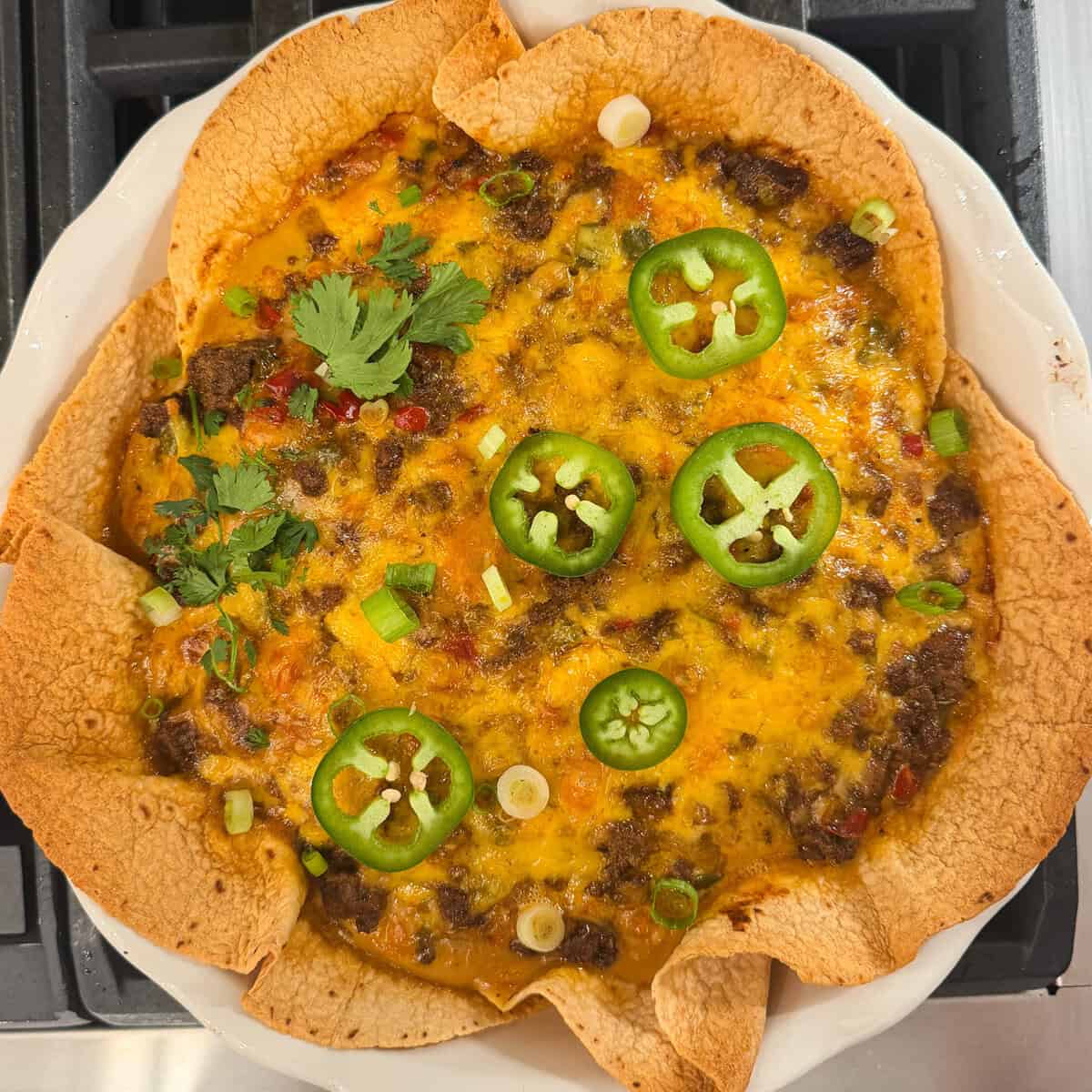 Taco pie with a crispy tortilla crust, melted cheese, ground beef, sliced jalapeños, cilantro, and green onions.