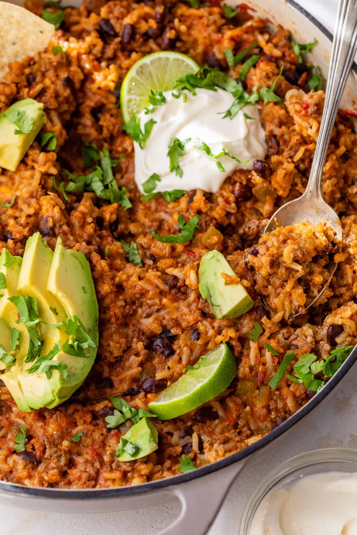 Cheesy taco rice with beans, avocado slices, lime wedges, sour cream, and fresh cilantro in a skillet with a spoon.