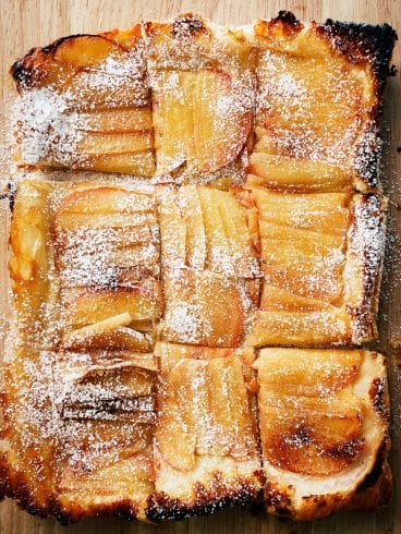 Rectangular upside down apple tart sliced into nine pieces, topped with powdered sugar, on a wooden surface.