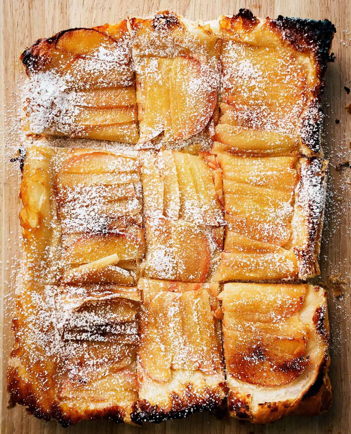 Rectangular upside down apple tart sliced into nine pieces, topped with powdered sugar, on a wooden surface.