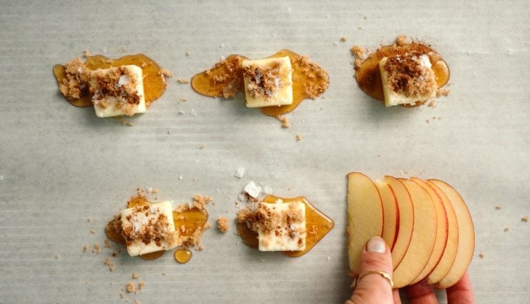 Five butter slices with honey and brown sugar on parchment; hand holding thin apple slices bottom right.