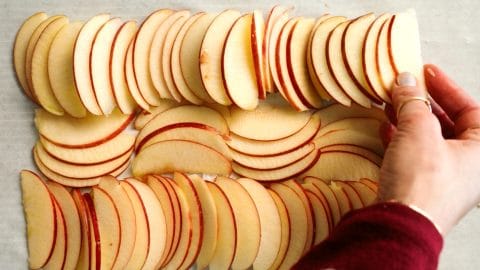 Hand arranging thinly sliced apple pieces in neat, overlapping rows on a white surface.