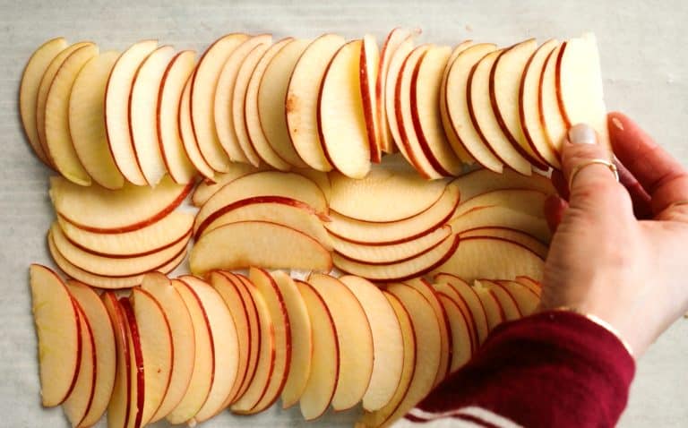 Hand arranging thinly sliced apple pieces in neat, overlapping rows on a white surface.