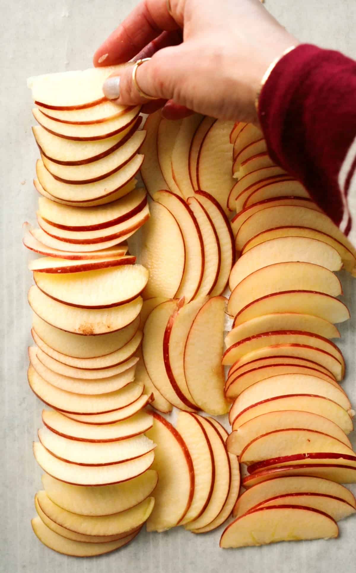 A hand arranges thinly sliced red apples into neat rows on a baking sheet